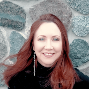 A smiling woman with red hair wearing a black top standing in front of a cobbled wall